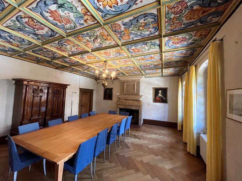 Ein grosser Holz-Esstisch mit blauen Stühlen steht in einem Raum mit buntem, bemaltem Deckenfresko. An der Wand sind zwei Gemälde und ein Kamin sichtbar.