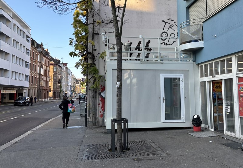 Strassenszene mit einem weissen Containergebäude neben einem Baum und einem Fussgänger, der eine Einkaufstasche trägt