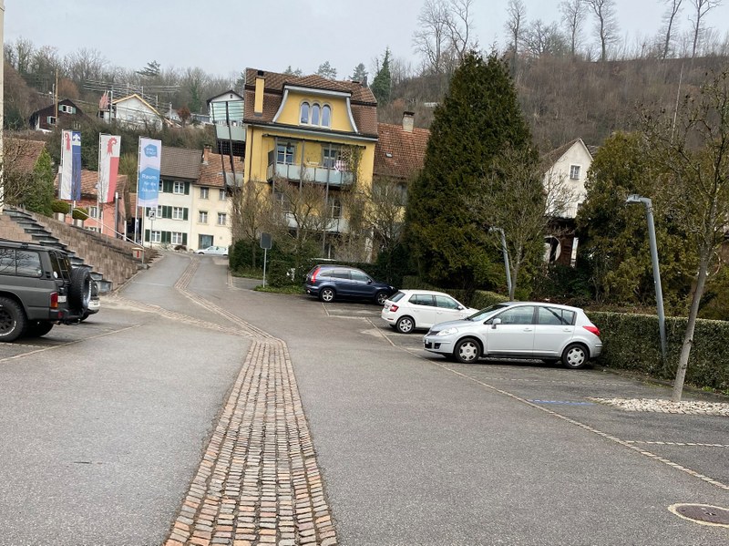 Parkplatz mit mehreren parkierten Autos vor Wohnhäusern und bewaldetem Hang im Hintergrund
