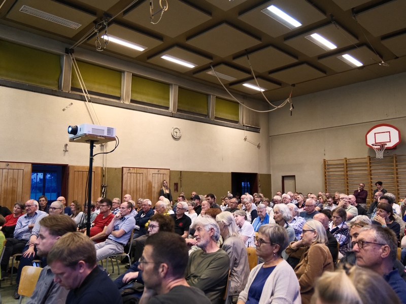 Grosse Gruppe von Menschen unterschiedlichen Alters sitzt in einer Turnhalle und hört aufmerksam zu, im Hintergrund Basketballkorb und Projektor