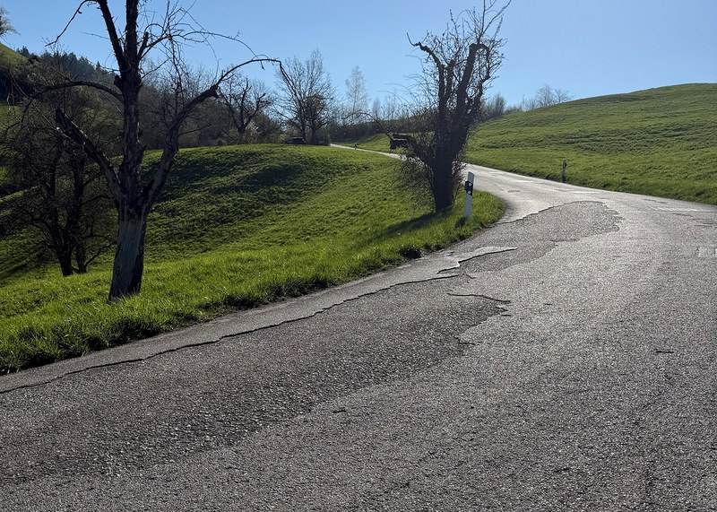 Landstrasse mit Schlaglöchern und Rissen, umgeben von grünen Hügeln und kahlen Bäumen unter klarem Himmel