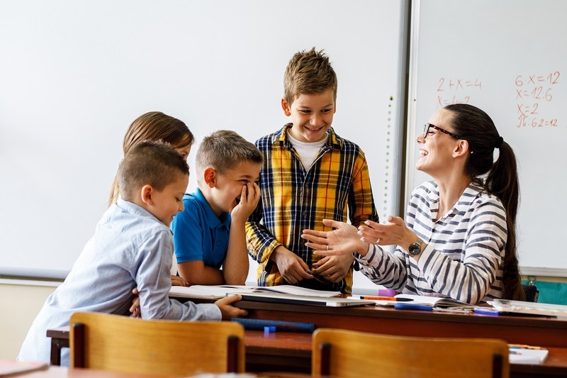 lachende Lehrerinn erklärt ihren ebenfalls lachenden 4 Schülern Matheaufgaben
