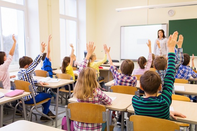 Primarschülerinnen und -schüler sind in einem Klassenzimmer mit ihrer Lehrerin zu sehen. Alle Kinder zeigen auf.