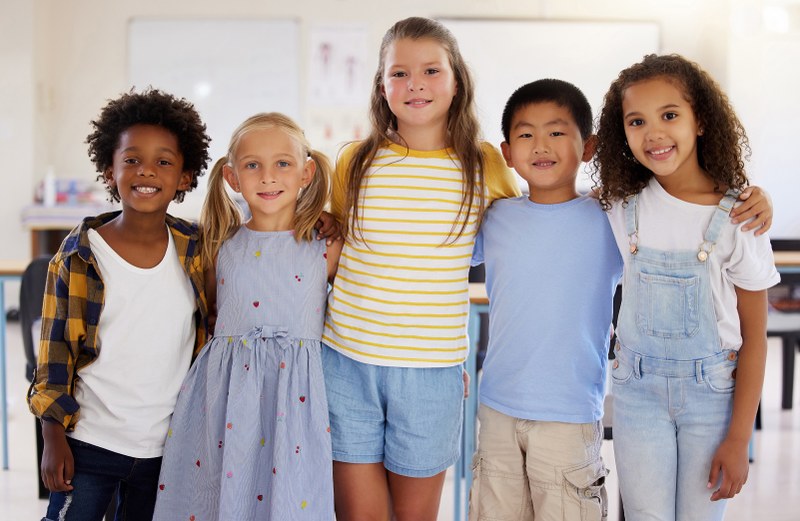 5 Schulkinder verschiedener Nationalitäten stehen in einem Klassenzimmer in einer Reihe nebeneinander und schauen direkt lächelnd in die Kamera