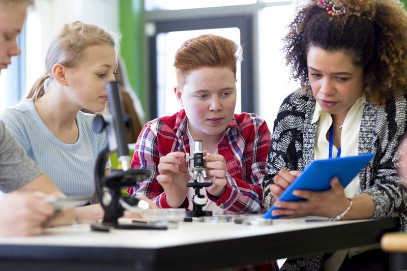 Eine Schülerin, ein Schüler beugen sich über ein Mikroskop, Lehrerin erklärt dazu etwas mit einem ipad in der Hand