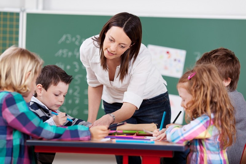 Lehrerin steht am Schülerpult und erklärt mehreren Schülerinnen und Schülern ihre Aufgaben
