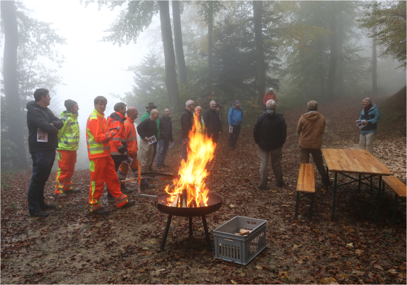 An der "Amtsübergabe" teilnehmende Personen stehen im nebligen Wald in grossem Kreis, im Vordergrund brennt in einer Schale ein grosses Feuer.
